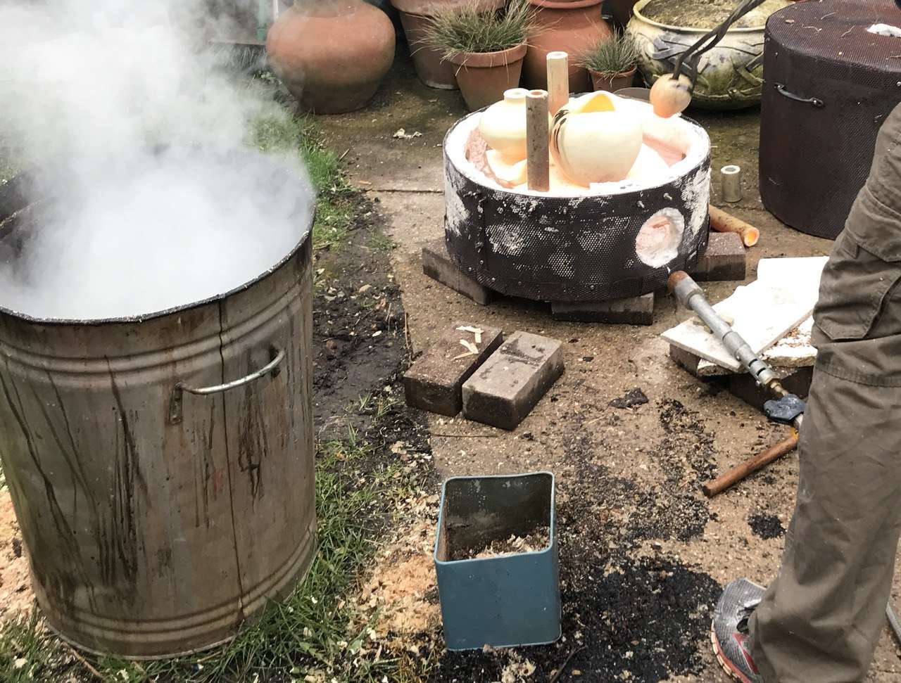 Piezas de cerámica calientes en una cocción raku, con humo y herramientas de trabajo alrededor.